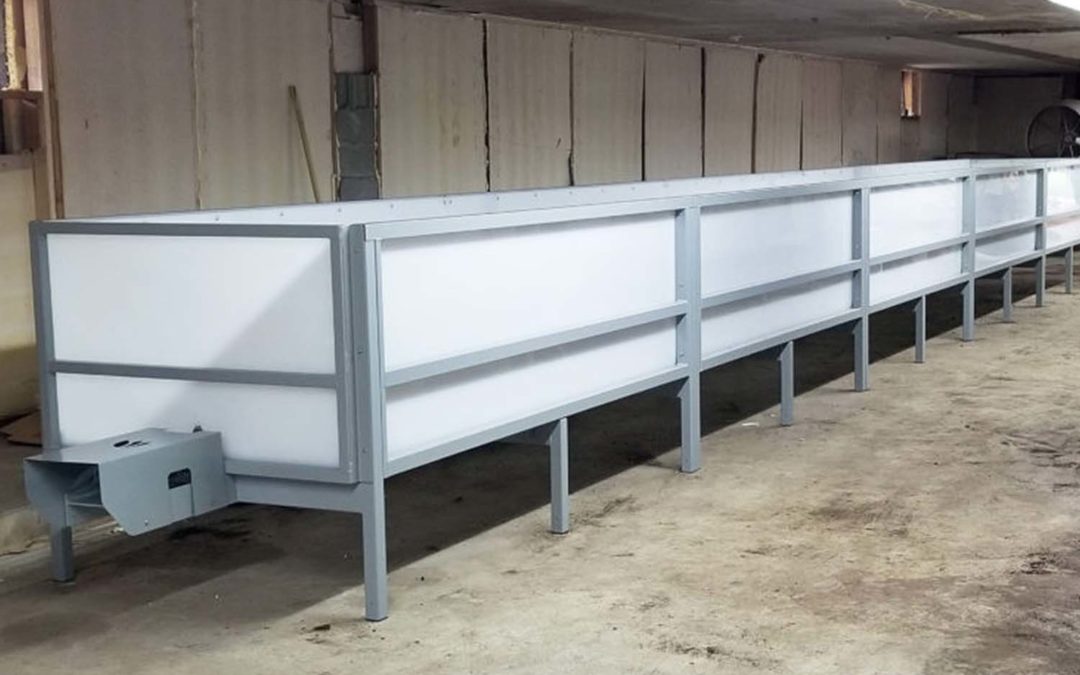 Image of a long, white continuous flow-through (CFT) composting system situated in a covered area with concrete flooring and wooden walls, representing a clean and professional setup for large-scale composting.